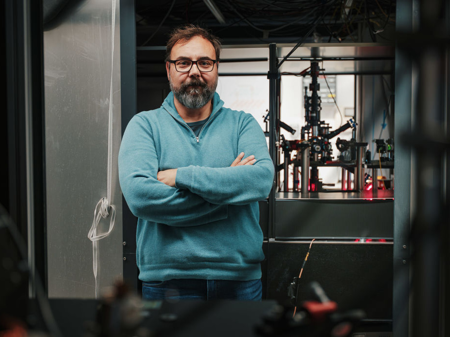 Giovanni Barontini, wearing glasses and a teal sweater, stands with his arms folded in front of the optical apparatus used to trap and cool rubidium atoms.