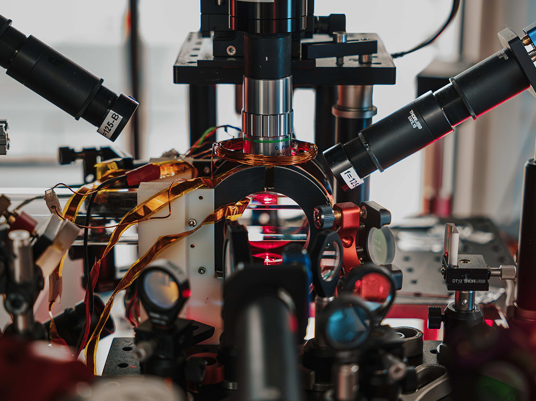 Close-up of the cold-atom optical table. Lenses and objectives surround a glass cell illuminated by red laser light, where a magneto-optical trap prepares a cloud of rubidium atoms.
