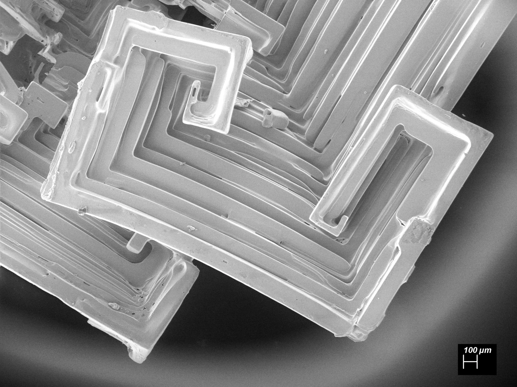 Greyscale scanning electron micrograph of potassium chloride crystals grown aboard the International Space Station. Nested square frames and stepped, hollow faces form scroll-like hopper structures; a 100-micrometre scale bar appears at lower right.