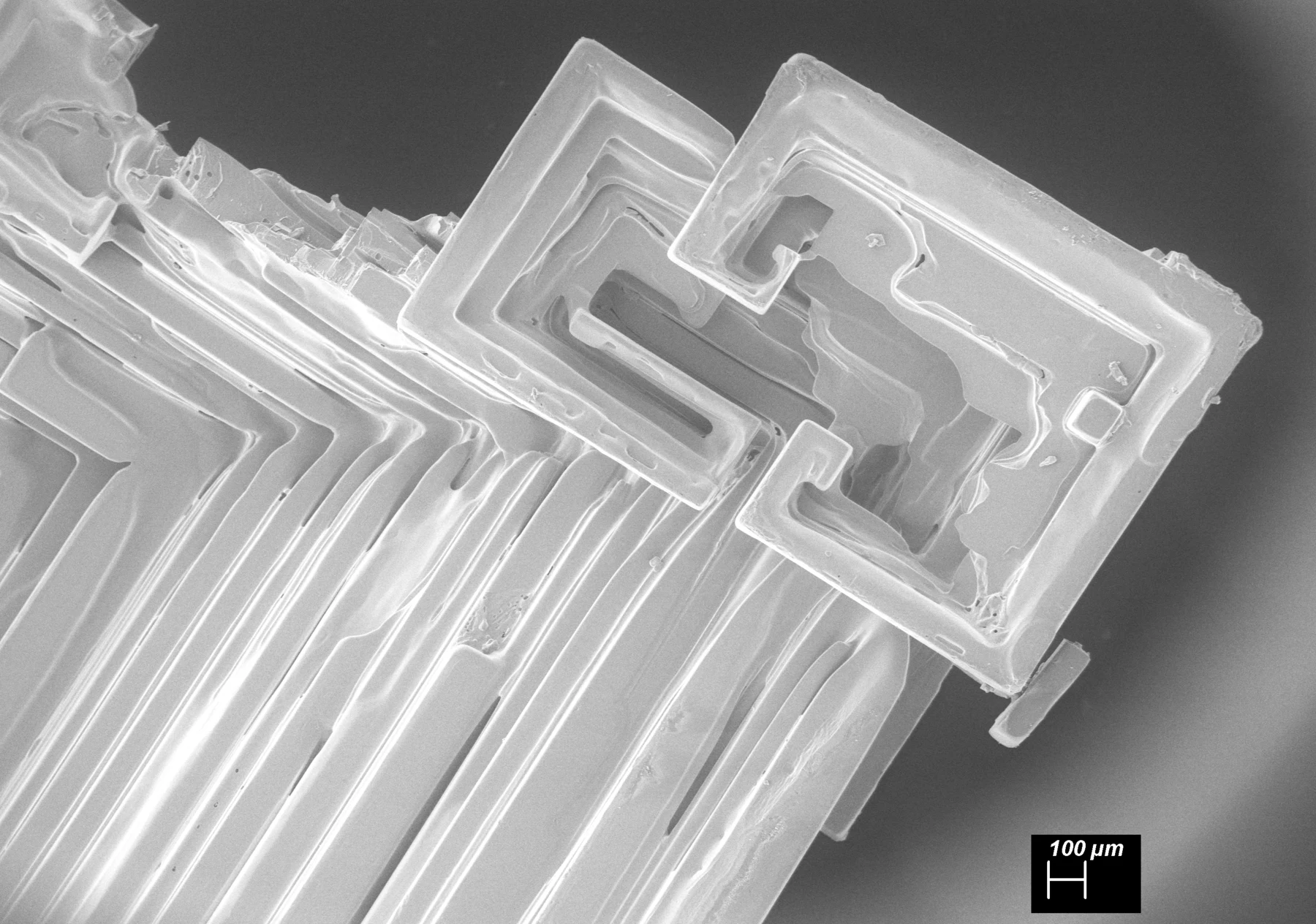 Greyscale scanning electron micrograph of a second potassium chloride scroll crystal grown aboard the International Space Station. Rectangular stepped sheets curl around one another like an inverted terraced structure; a 100-micrometre scale bar appears at lower right.